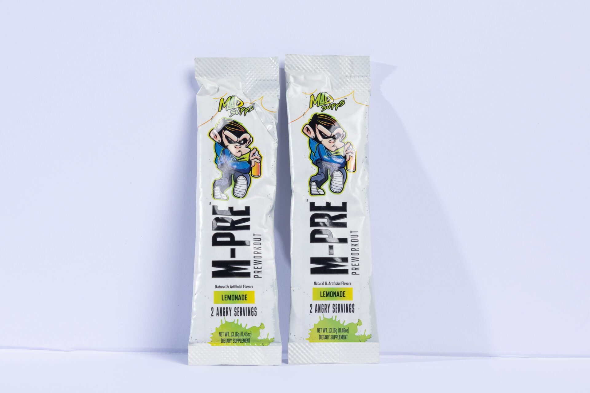 Two white pre-workout packets with a cartoon monkey logo, lemonade flavor, standing upright on a pastel purple surface.