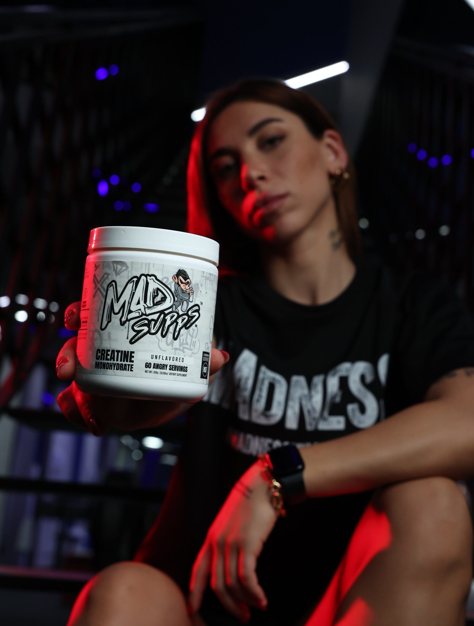 Tub of Mad Supps creatine monohydrate held up in foreground, label visible, person blurred in dark venue with red lighting behind.