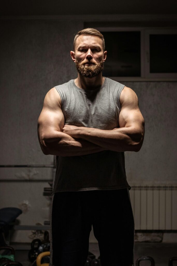 Muscular man with a beard stands in a dim gym, arms crossed, wearing a gray sleeveless shirt.