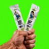 Two white sachets with a cartoon character held in a clenched fist against a bright neon-green background, showing a bold athletic design and flavor labels at the bottom.